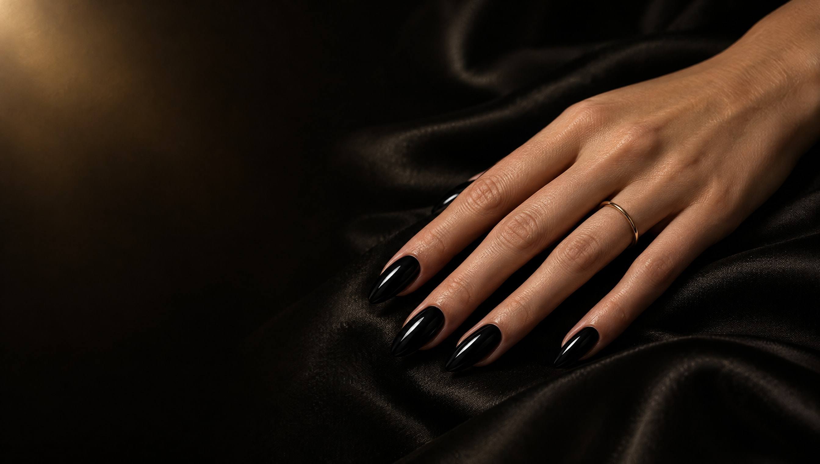 Hand with long glossy black almond nails and a thin gold ring resting on black silk